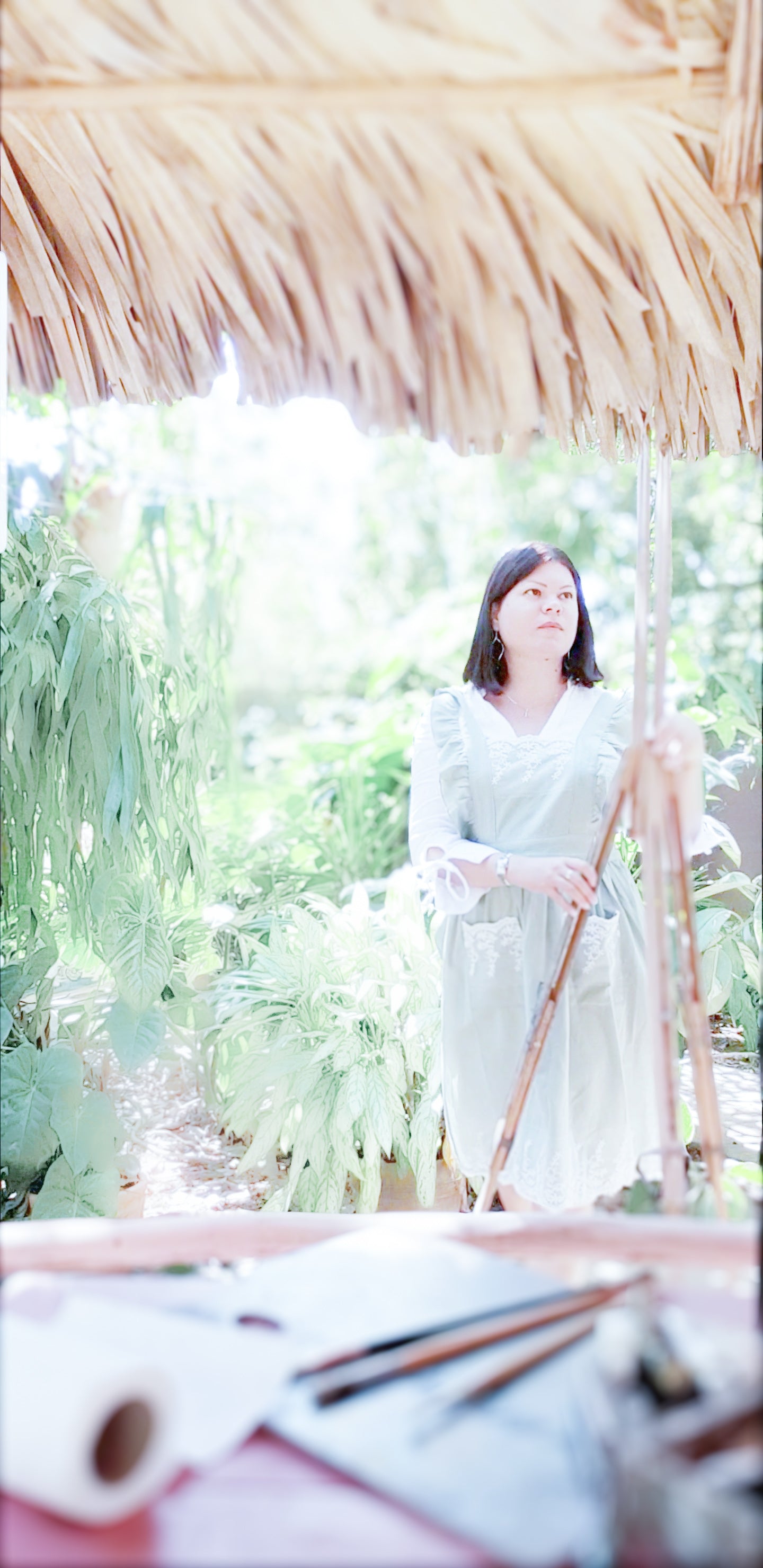 ARTIST Woman in a light dress standing holding a easel under a thatched roof with greenery in the background